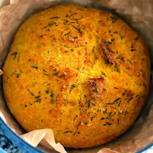 Square photo of a loaf of pumpkin no knead bread in a parchment lined Dutch oven, with rosemary and sea salt on top.