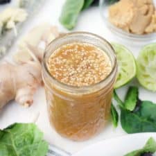Close-up of open glass jar filled with peanut dressing with sesame seeds mixed in, on white surface with fresh produce scattered around the jar.