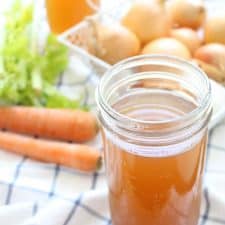 Glass jar full of vegetable stock on a blue and white checkered tablecloth, with vegetables in the background.