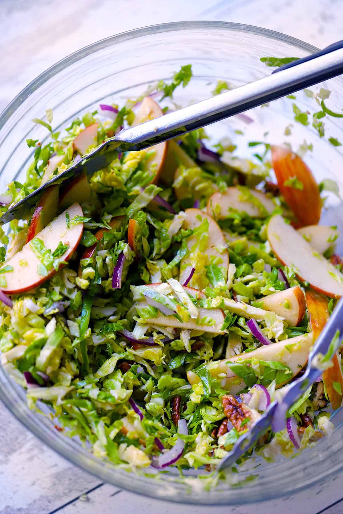 A shaved brussels sprouts salad being tossed in a clear bowl with apples, dressing, onions, and candied pecans.