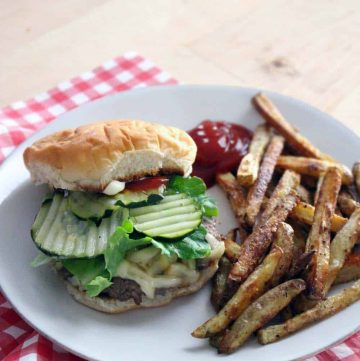 Cheeseburger with pickles, ketchup, and lettuce on a white plate with french fries and a dollop of ketchup, on a red and white checkered tablecloth.