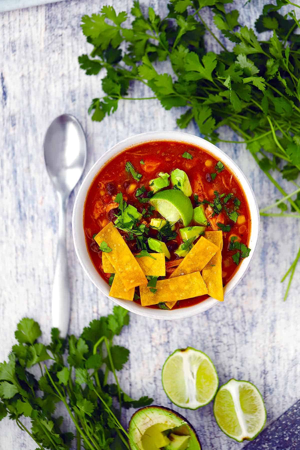 Overhead photo of a bowl of chicken tortilla soup surrounded by cilantro and limes.