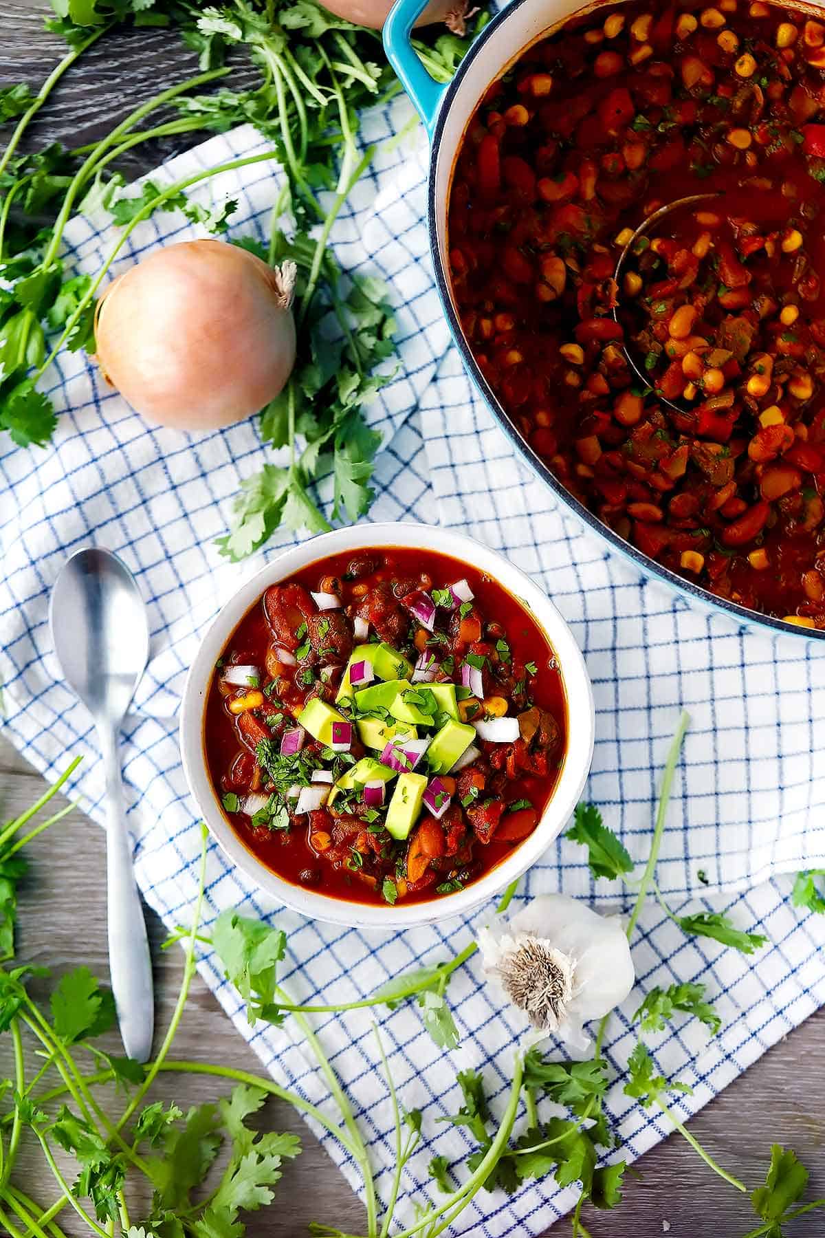 A flatlay photo of vegetarian chili with a dutch oven of chili next to it and cilantro and onions and a towel in the background.