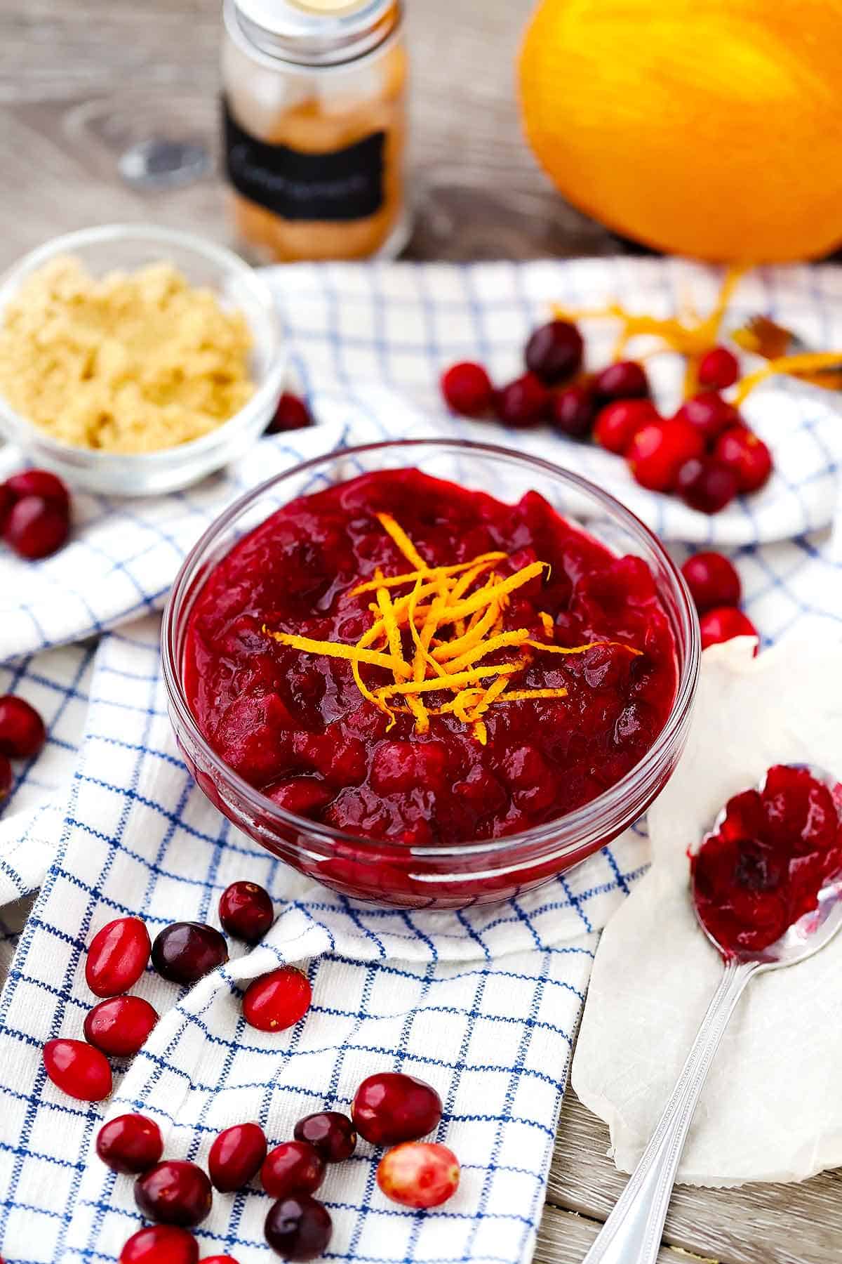 A bowl of fresh whole cranberry sauce with orange zest on top and fresh cranberries, brown sugar, and cinnamon in the background.