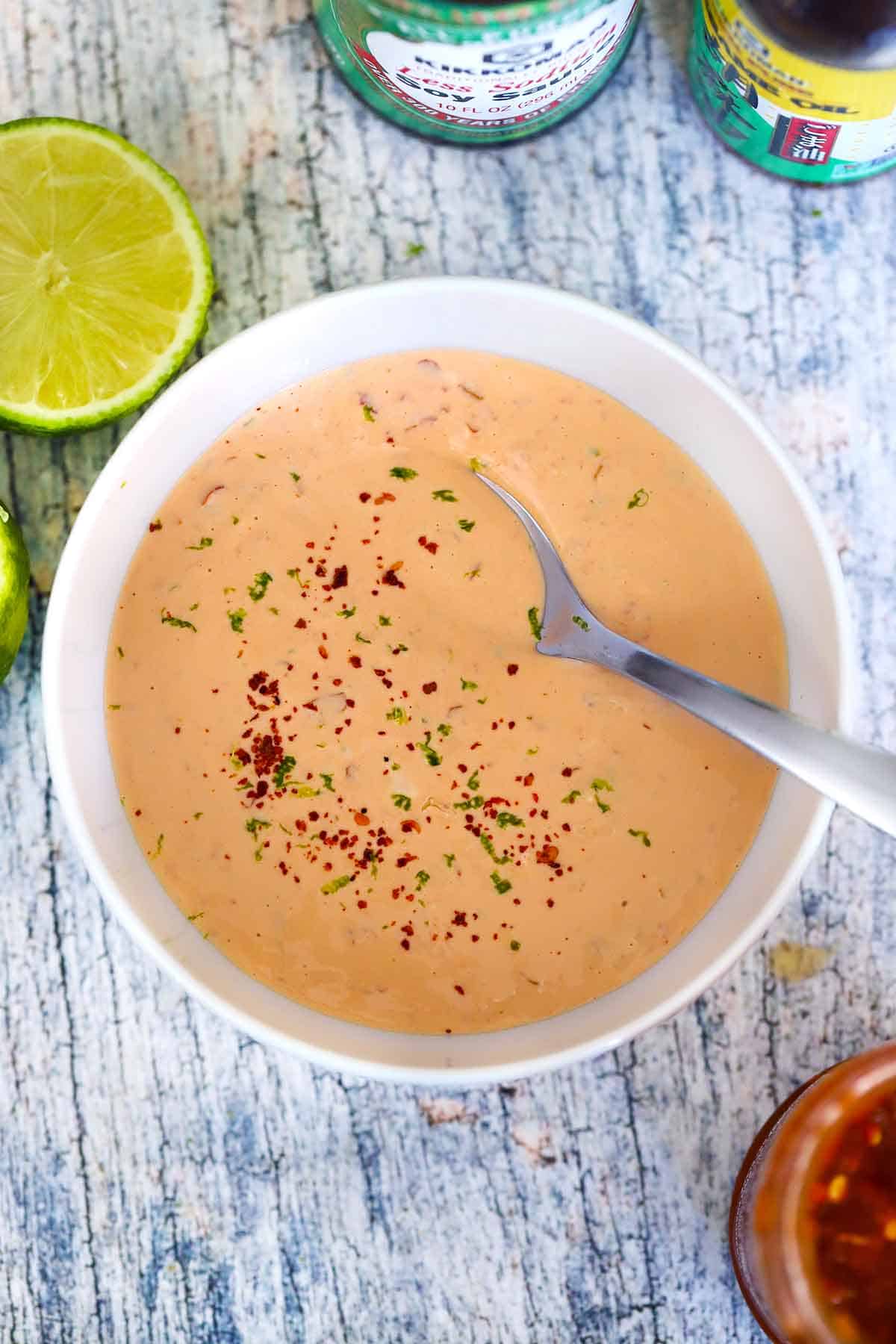 Overhead view of a bowl of homemade spicy mayo with a spoon in it, topped with lime zest and red pepper.