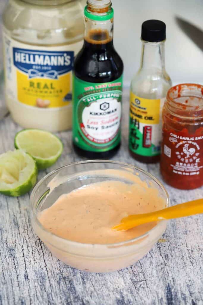A bowl of spicy mayo in the foreground with mayonnaise, a lime, soy sauce, toasted sesame oil, and chili garlic sauce displayed in the background.
