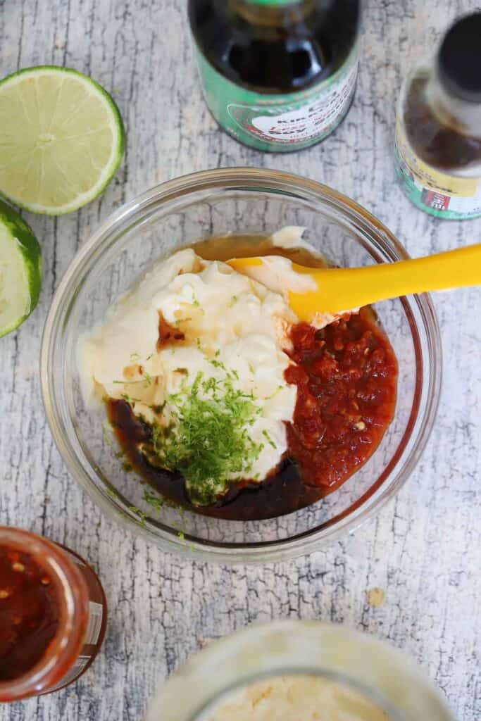 A clear bowl with the ingredients for spicy mayo added to it and displayed around the bowl, including mayonnaise, chili garlic sauce, soy sauce, sesame oil, and lime.