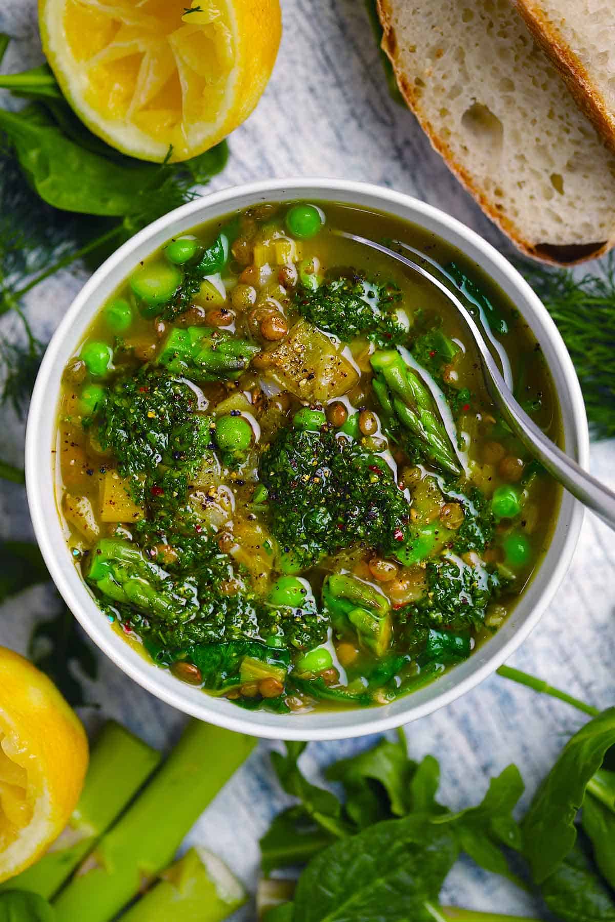 Overhead view of a bowl of spring lentil vegetable soup with bright green veggies and herb pistou spooned on top with ingredients like lemon, asparagus, and spinach scattered around it.