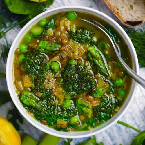 Square photo of an overhead view of spring lentil vegetable soup with bright green veggies and herb pistou spooned on top.