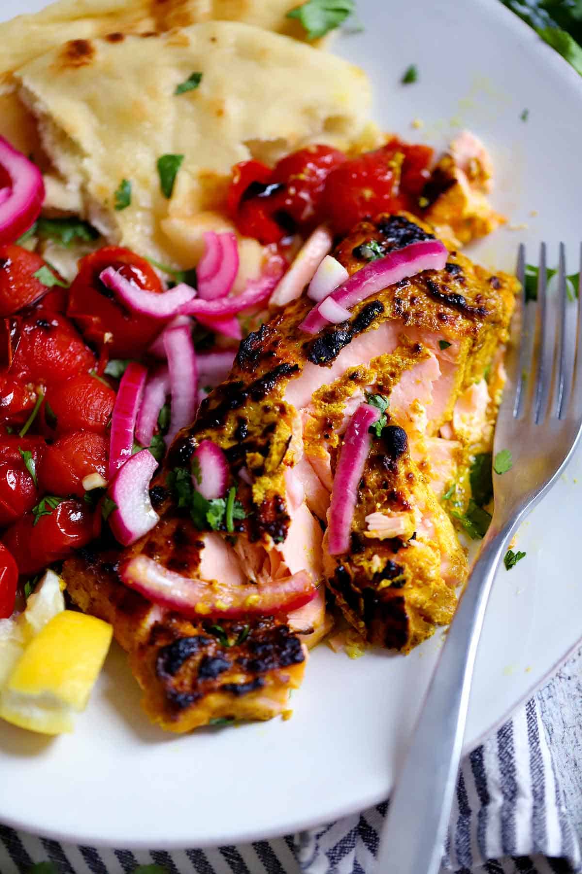 A plate with tandoori salmon that's been flaked apart with cherry tomatoes, topped with pickled red onions and a fork alongside.