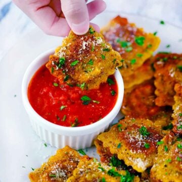 Square photo of a hand dipping a toasted ravioli into marinara sauce.