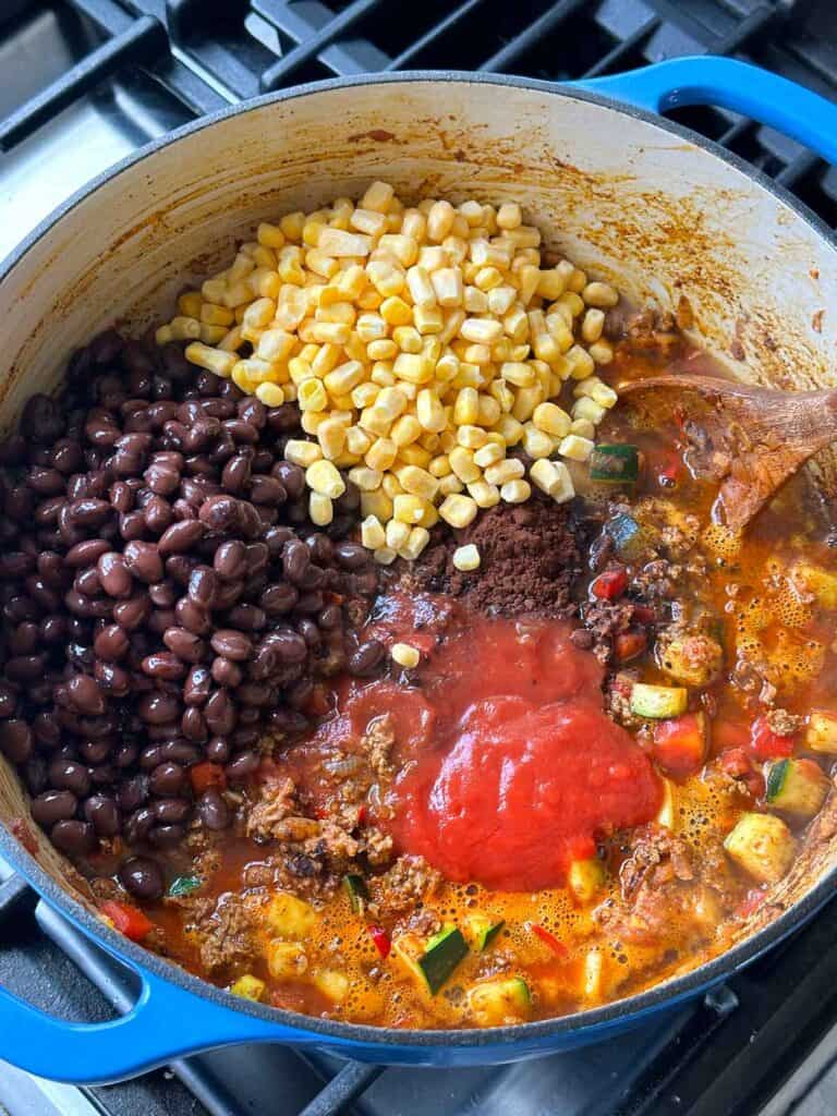 Adding black beans, corn, and crushed tomatoes to turkey chili in a Dutch oven.