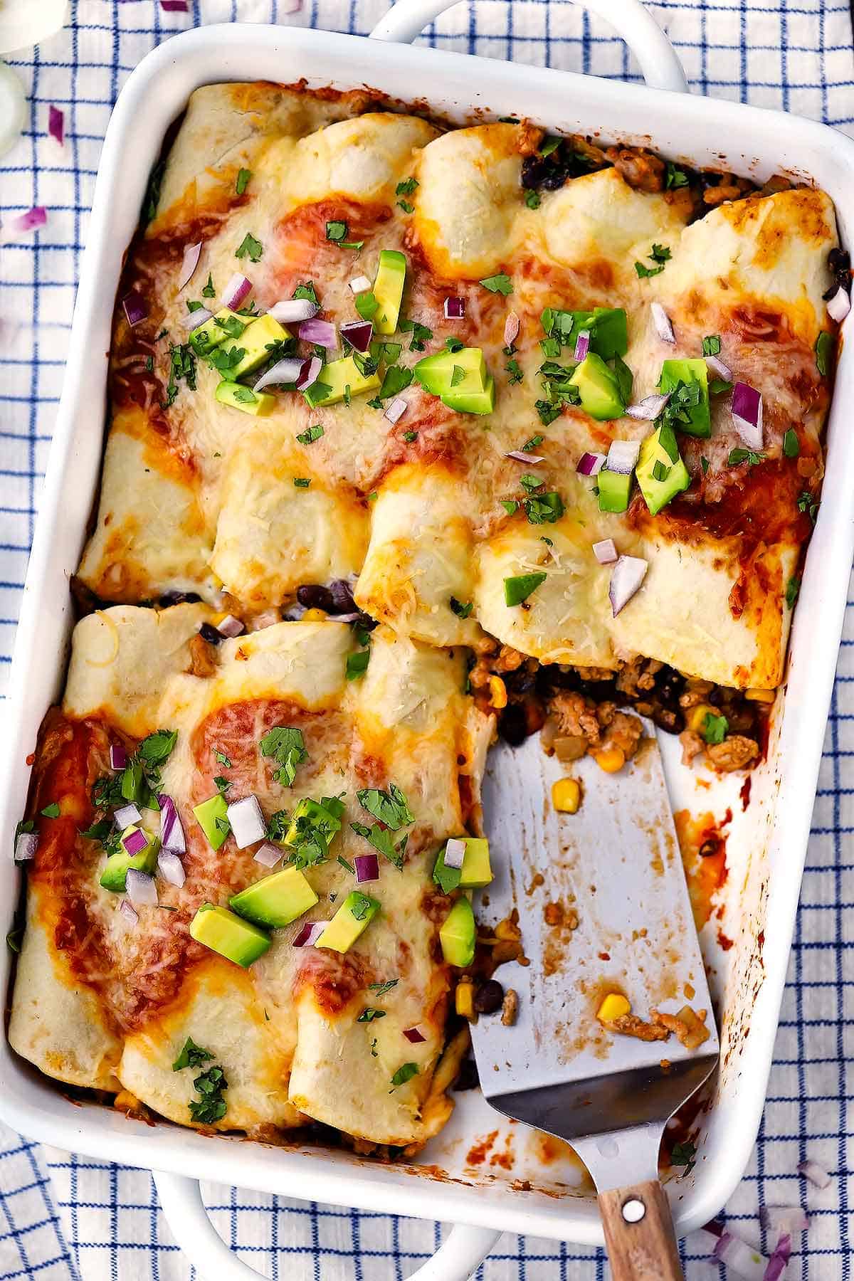 A baking dish with turkey enchiladas with black beans and corn and a spatula.