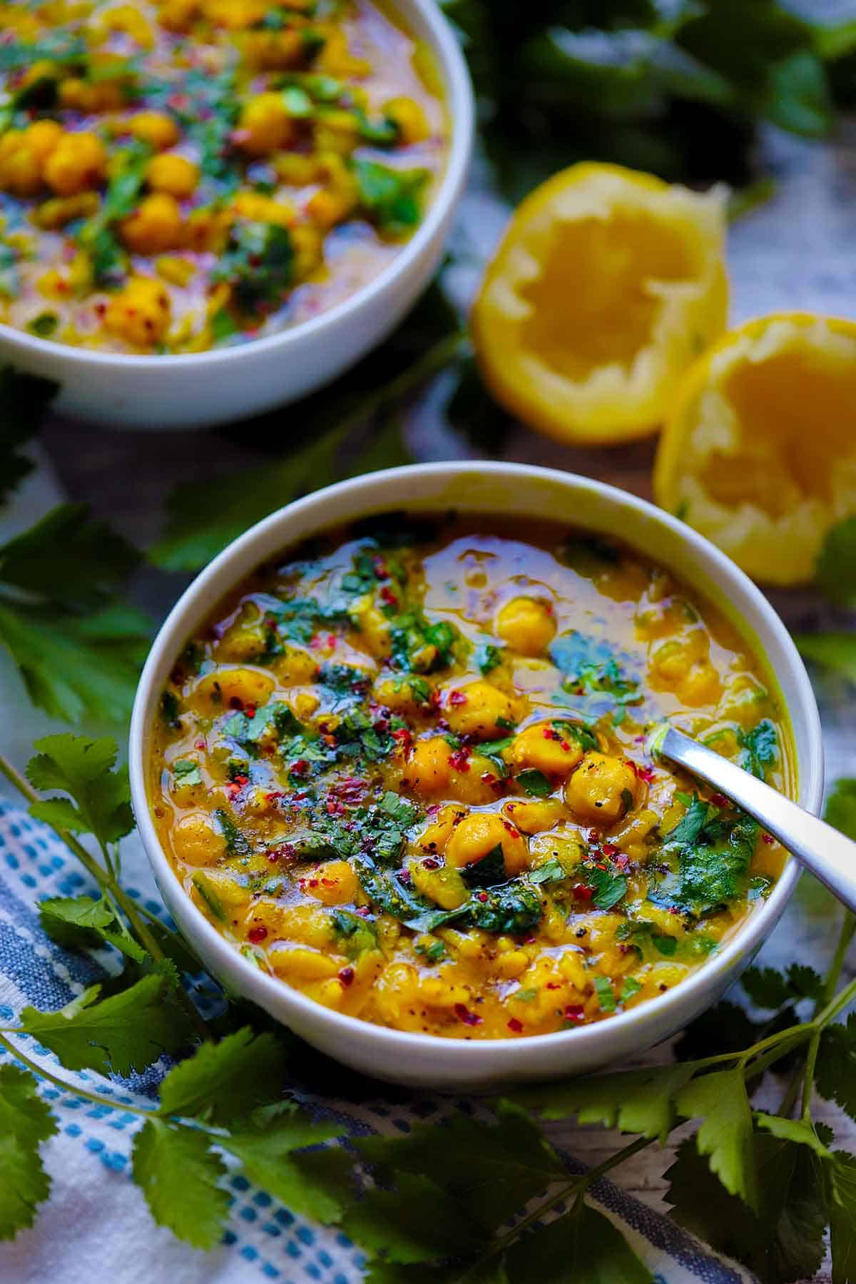 Side view of two bowls of chickpea, orzo, and spinach soup with turmeric and lemon, with a spoon in one bowl, with parsley and lemon scraps scattered around.
