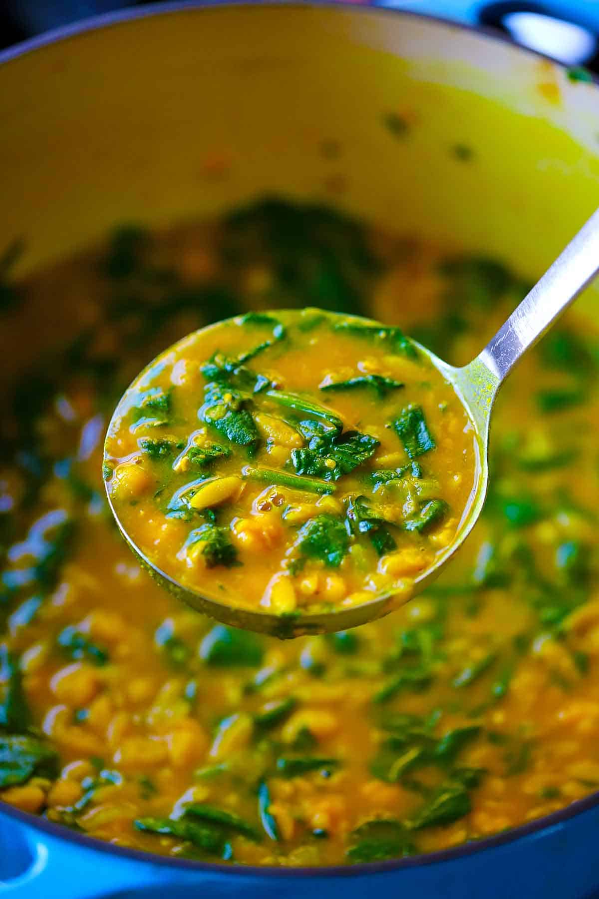A ladle with turmeric soup over a blue Dutch oven pot.