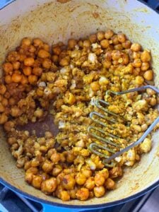 Using a potato masher to mash chickpeas after sautéing in spices in a pot.