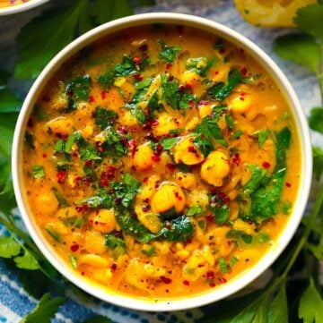 Square photo of overhead view of a bowl of turmeric soup with orzo, chickpeas, spinach, and lemon, with aleppo pepper and olive oil on top.