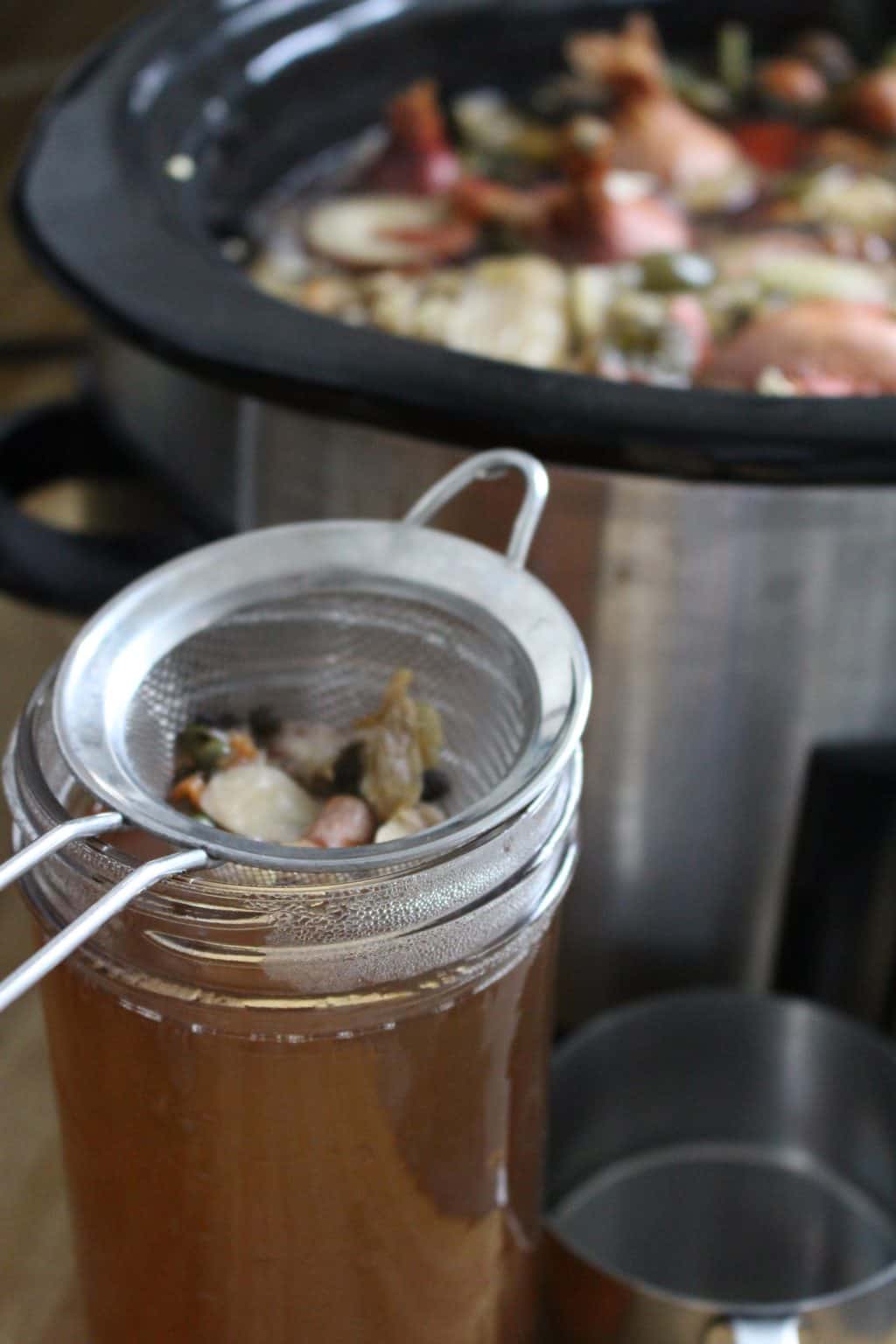Small mesh strainer placed over opening of mason jar, with small vegetable scraps in the strainer and vegetable broth in the jar.